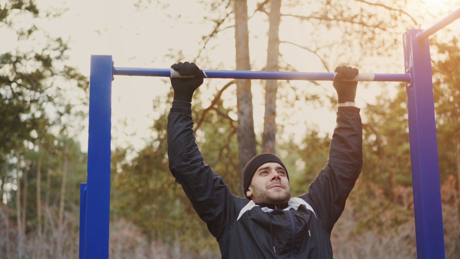 A man hanging from a blue outdoor pull-up bar in a winter park, wearing gloves and a beanie